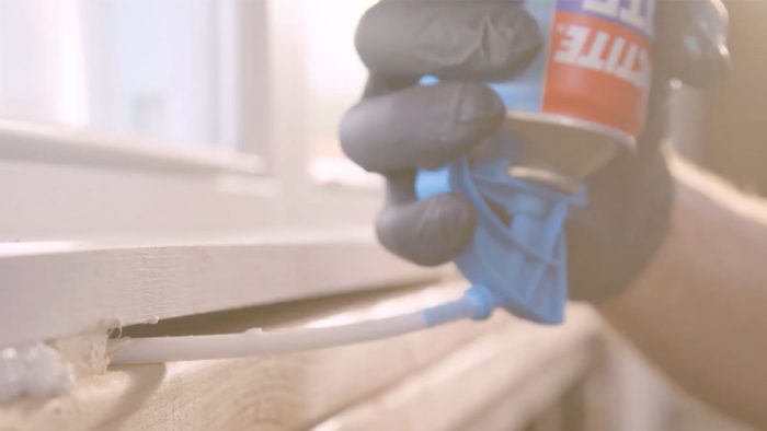 A close up of a hand wearing black rubber gloves using a bottle of Loctite TITE FOAM Window & Door to air seal the area.