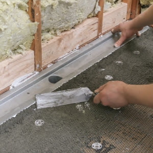 I'm using a Laticrete linear drain here, setting it in the dry-pack mud-bed shower pan before installing the bottom piece of foamboard over its back edge.